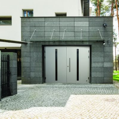 Moderni harmaa autotalli, jossa on suuri kaksinkertainen nosto-ovet, jossa on pystysuorat lasipaneelit. Rakennuksessa on kaakeloitu ulkopinta, ja sitä ympäröi mukulakivipäällyste, puita ja nurmikkoinen piha.